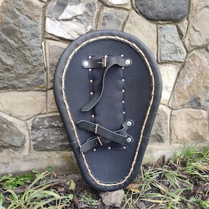 May include: Black leather shield with a teardrop shape, featuring a stitched border and riveted details. The back has leather straps for securing the shield. The shield is set against a stone wall.