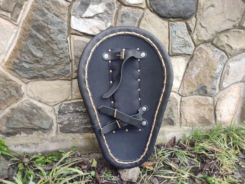 May include: Black leather shield with a teardrop shape, featuring a stitched border and riveted details. The back has leather straps for securing the shield. The shield is set against a stone wall.