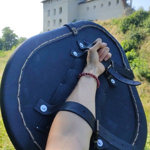 May include: A black, round shield with leather straps and a rope-stitched edge. The shield is held up against a backdrop of a stone castle and green foliage.
