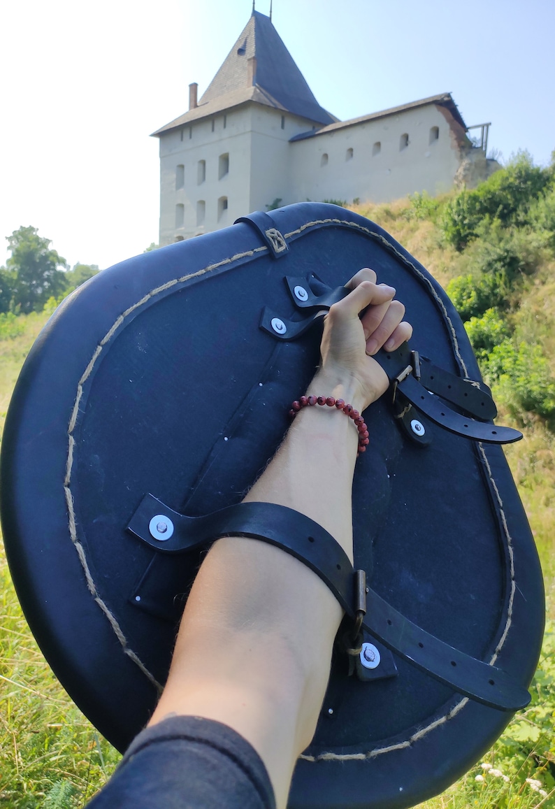 May include: A black, round shield with leather straps and a rope-stitched edge. The shield is held up against a backdrop of a stone castle and green foliage.