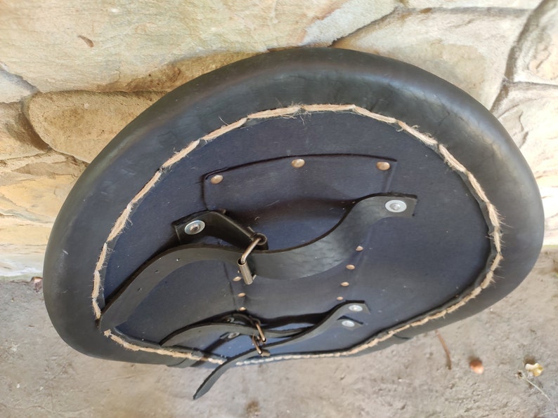 May include: A black leather shield with a rounded shape, featuring a dark blue interior and a stitched border. The shield has leather straps and metal hardware for securing it. The shield is likely used for protection.