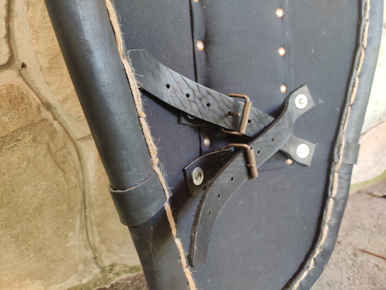 May include: Close-up of a black leather shield with riveted accents and leather straps. The shield has a curved shape and is edged with a lighter-colored material. The straps are secured with metal buckles.