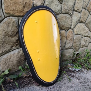 May include: A yellow, oval-shaped shield with a black border and decorative rivets. The shield is set against a stone wall, suggesting a historical or medieval theme. The bright yellow color is eye-catching.
