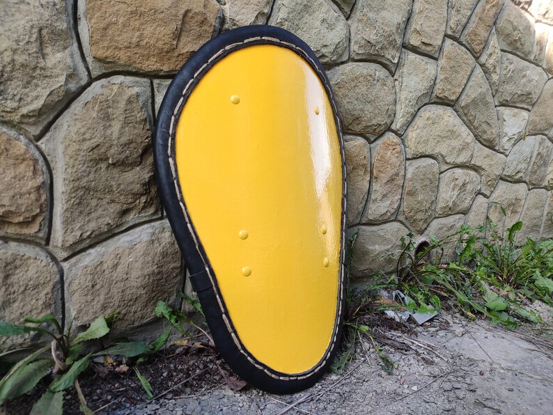 May include: A yellow, oval-shaped shield with a black border and decorative rivets. The shield is set against a stone wall, suggesting a historical or medieval theme. The bright yellow color is eye-catching.