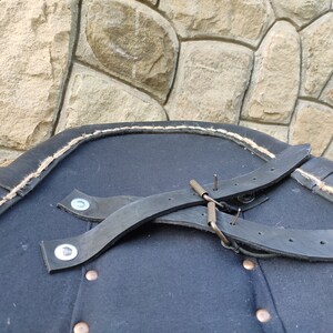 May include: A close-up of a dark blue garment with black leather straps and metal hardware. The edge is trimmed with a light-colored rope. The garment has several copper-colored buttons.