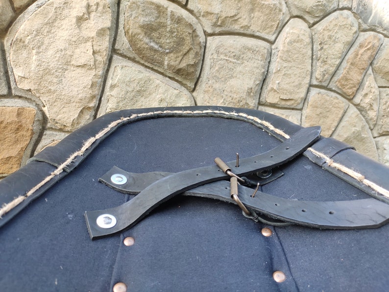 May include: A close-up of a dark blue garment with black leather straps and metal hardware. The edge is trimmed with a light-colored rope. The garment has several copper-colored buttons.