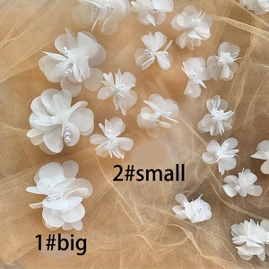 May include: A collection of white fabric flowers with pearl accents. There are two sizes of flowers, with one large flower and two smaller flowers. The flowers are arranged on a beige mesh fabric.