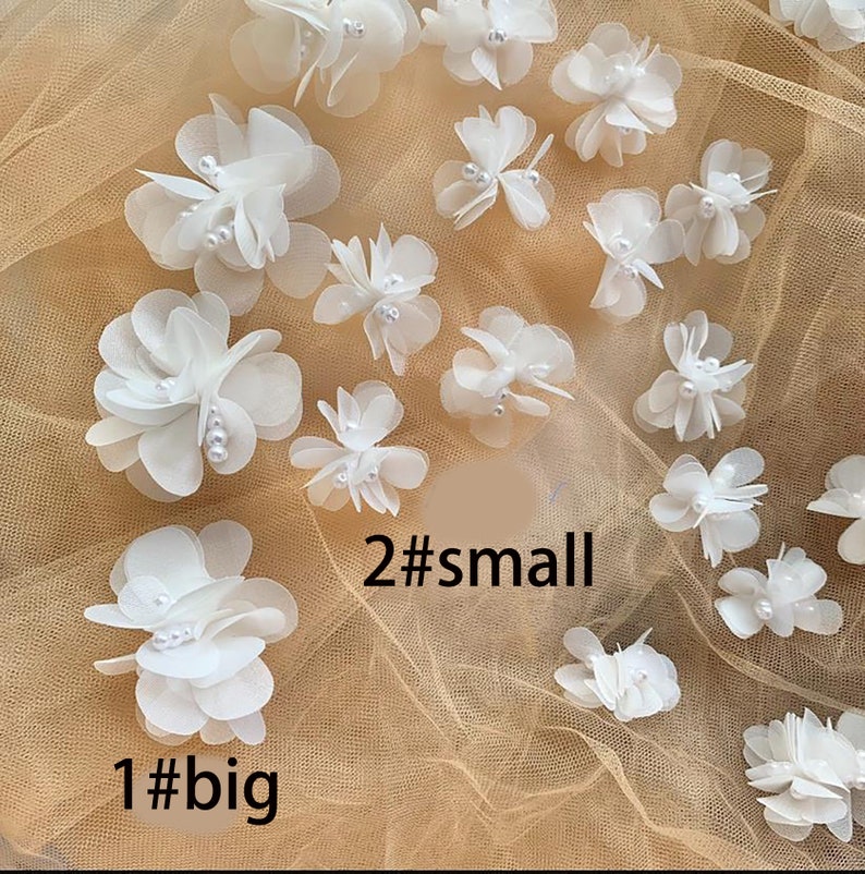 May include: A collection of white fabric flowers with pearl accents. There are two sizes of flowers, with one large flower and two smaller flowers. The flowers are arranged on a beige mesh fabric.