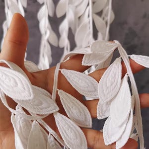 Pode incluir: Renda branca com um design de folha. A renda é feita de tecido delicado e tem uma textura macia e fluida.