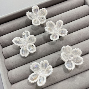 Może przedstawiać: Pięć białych koralikowych kwiatów z przezroczystymi kryształowymi środkami. Kwiaty są ułożone na szarej powierzchni.