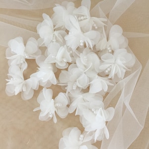 May include: A collection of white fabric flowers with pearl accents. The flowers are arranged on a white tulle fabric background.