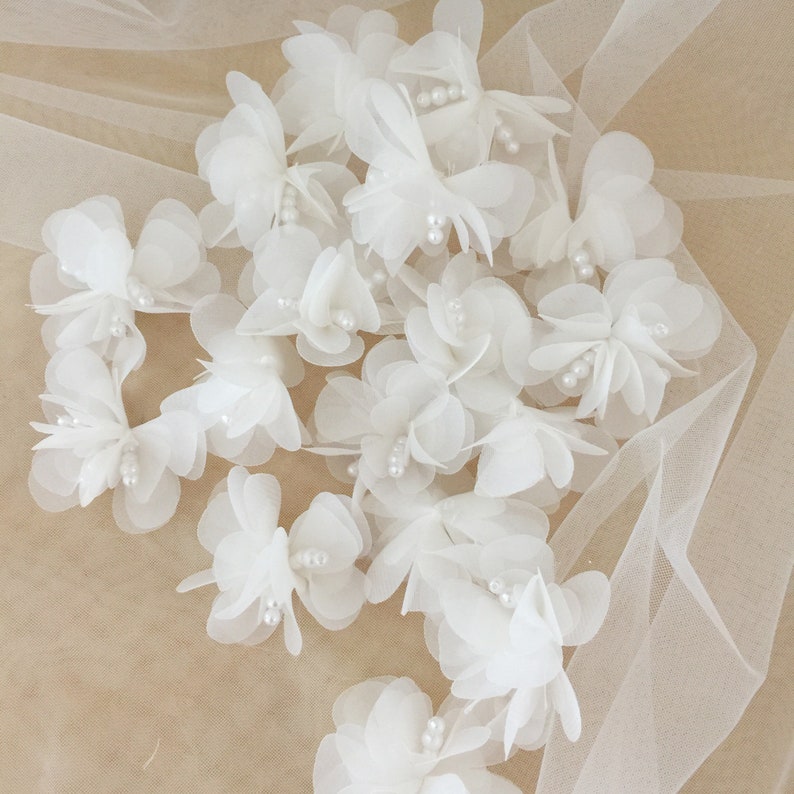 May include: A collection of white fabric flowers with pearl accents. The flowers are arranged on a white tulle fabric background.