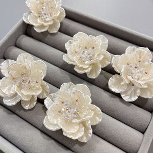 Puede incluir: Cinco adornos en forma de flor blanca con detalles dorados y brillantes de cristal transparente. Los adornos se muestran en una caja de terciopelo gris.