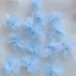 May include: A collection of light blue fabric flower embellishments. The flowers are made of multiple layers of sheer fabric and have a delicate, ruffled appearance.