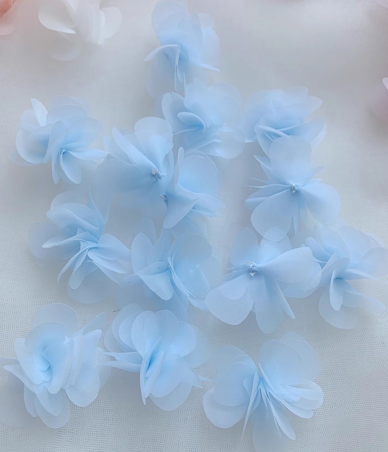 May include: A collection of light blue fabric flower embellishments. The flowers are made of multiple layers of sheer fabric and have a delicate, ruffled appearance.