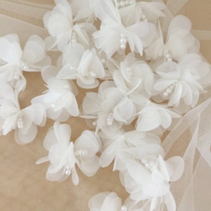 May include: A collection of white fabric flowers with pearl accents. The flowers are arranged on a white tulle background.