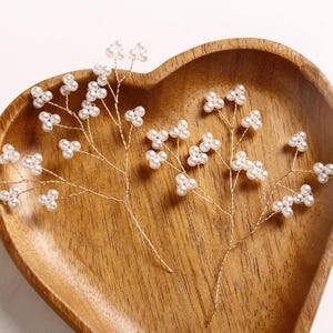 May include: Close-up of two delicate, gold-wired hair accessories, each adorned with clusters of small, white pearls. The accessories are arranged on a heart-shaped wooden tray, creating a natural and elegant aesthetic. Ideal for bridal or special occasion hairstyles.