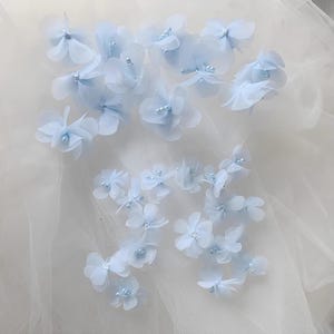 May include: A collection of light blue fabric flowers with pearl accents. The flowers are scattered on a white tulle background.
