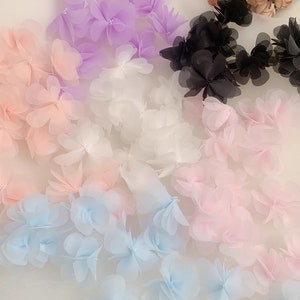 May include: Assortment of delicate flower-shaped trims in various colours, including light pink, white, light blue, lavender, and black. The trims are made of sheer fabric and have a soft, feminine look.