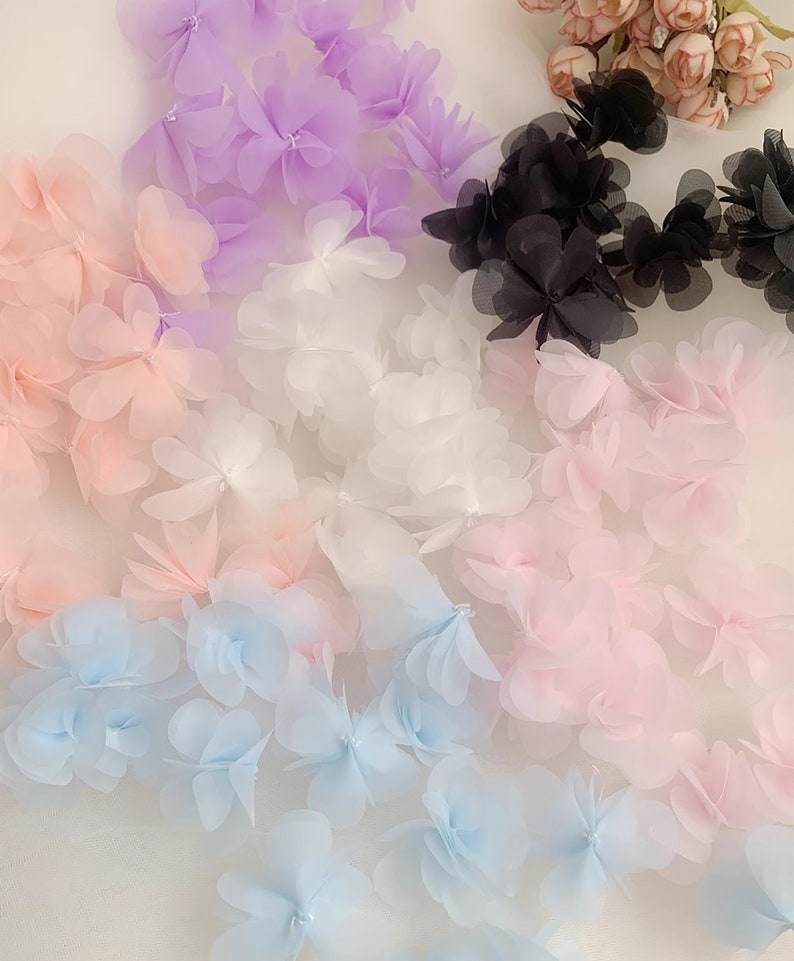 May include: Assortment of delicate flower-shaped trims in various colours, including light pink, white, light blue, lavender, and black. The trims are made of sheer fabric and have a soft, feminine look.