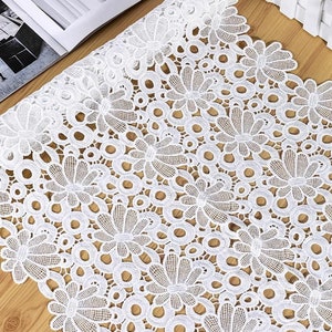 Pode incluir: Tecido de renda branca com um delicado padrão floral. A renda apresenta um desenho repetitivo de pequenas flores e círculos.