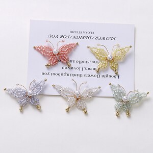 Pode incluir: Cinco broches de borboleta com contas em cores pastel: rosa, amarelo, lavanda, branco e verde. Cada borboleta tem um corpo de pérola e detalhes dourados. As borboletas estão dispostas em um cartão branco com texto.