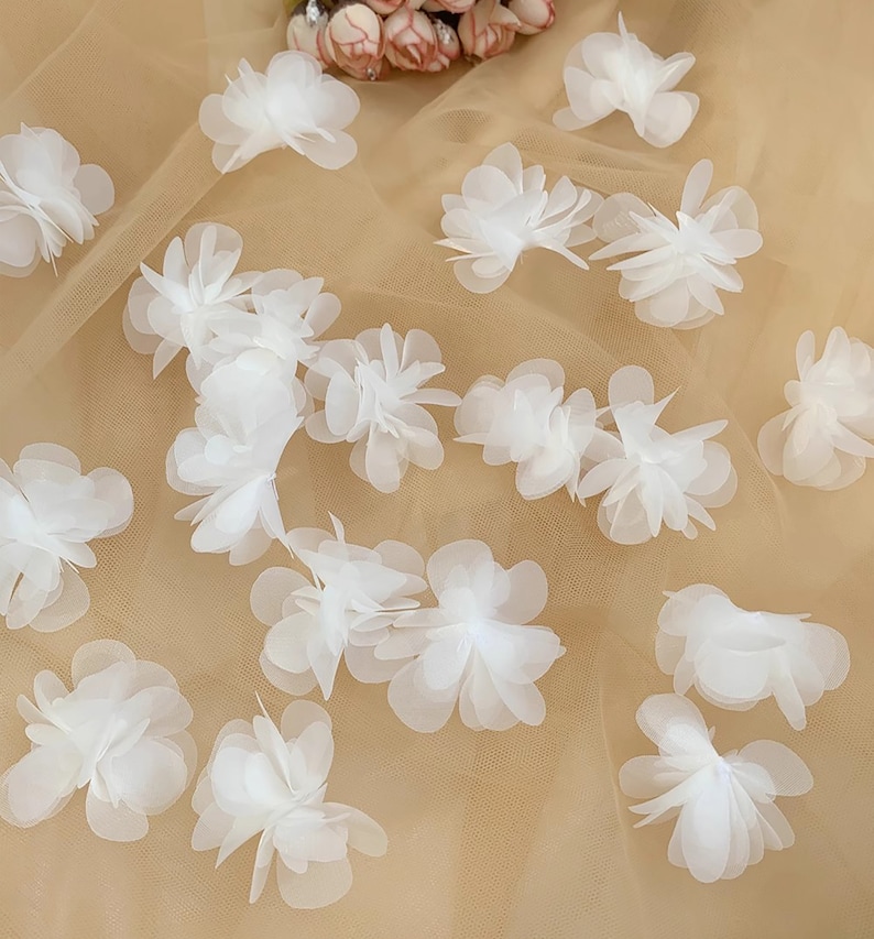 May include: White fabric flower embellishments on a beige mesh background. The flowers are layered and have a delicate, ruffled appearance.