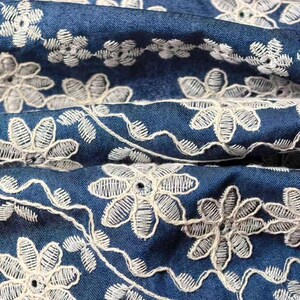 May include: Close-up of blue denim fabric with white floral embroidery. The embroidery features detailed flower and leaf patterns, creating a delicate and textured design. The fabric is folded, showing the depth and texture of the material.