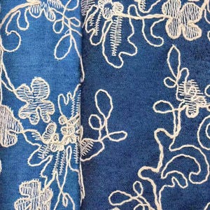 May include: Close-up of blue fabric with white floral embroidery. The embroidery features detailed flower and leaf patterns. The fabric has a textured appearance, and the stitching is intricate, creating a delicate design. The overall effect is a blend of texture and pattern.