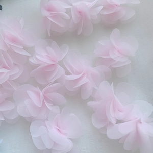May include: Pink fabric flower embellishments on a white mesh background. The flowers are layered and have a delicate, 3D appearance.