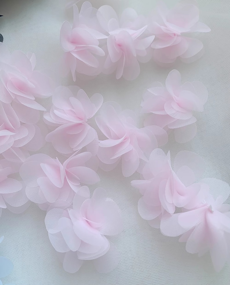 May include: Pink fabric flower embellishments on a white mesh background. The flowers are layered and have a delicate, 3D appearance.
