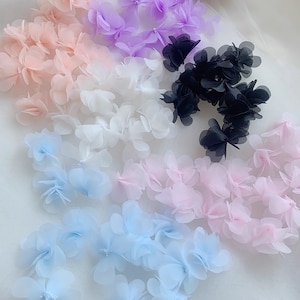 May include: An assortment of delicate, layered fabric flowers in various colours, including pink, white, light blue, purple, and black. The flowers are arranged in rows on a white background.