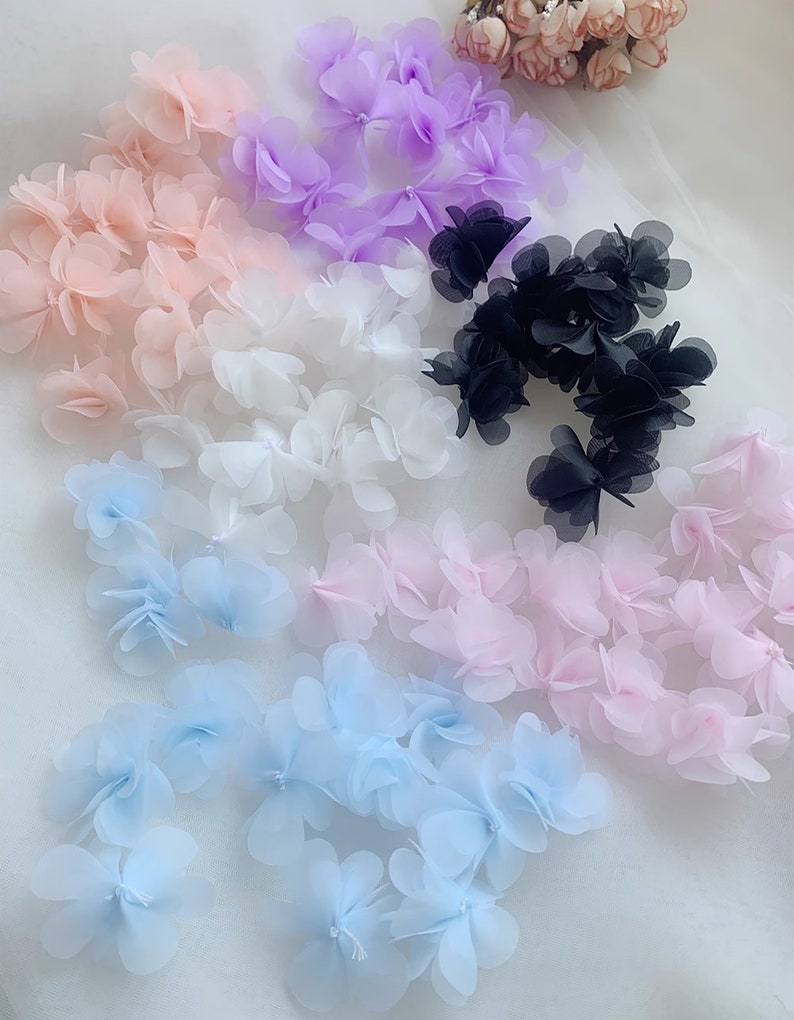 May include: An assortment of delicate, layered fabric flowers in various colours, including pink, white, light blue, purple, and black. The flowers are arranged in rows on a white background.