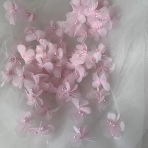 May include: A collection of pink fabric flowers with pearl centers. The flowers are scattered on a white tulle background.