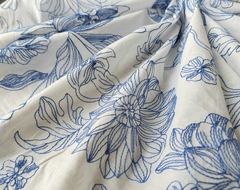 May include: White fabric with a blue floral embroidery pattern. The fabric is folded and shows the intricate details of the design.