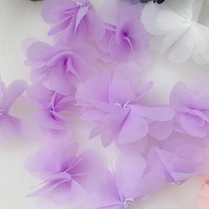 May include: A collection of lavender-coloured fabric flowers. The flowers are made of a sheer, delicate material and have a layered, ruffled appearance.