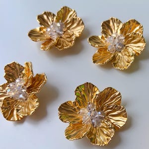 Könnte beinhalten: Vier goldfarbene, blütenförmige Verzierungen mit strukturierten Blütenblättern und einem Cluster aus klaren Kristallen in der Mitte. Das florale Design hat ein glänzendes Finish. Diese dekorativen Elemente könnten für Schmuck- oder Bastelprojekte verwendet werden.