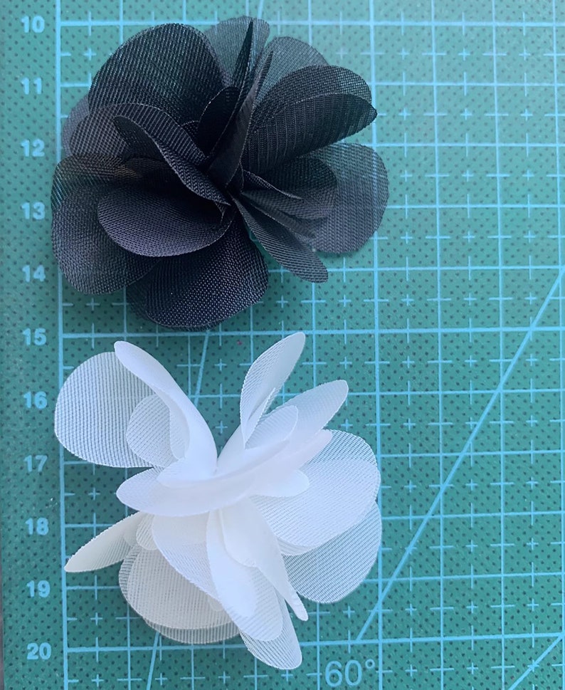 May include: Two fabric flowers, one black and one white, on a green cutting mat. The flowers are made of multiple layers of fabric and have a ruffled appearance.