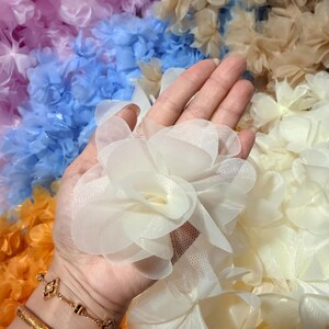 Può includere: Primo piano di una mano che tiene un gruppo di fiori di stoffa bianchi. I fiori hanno più strati di petali delicati. Lo sfondo presenta una varietà di fiori di stoffa colorati nelle tonalità del blu, arancione e beige.