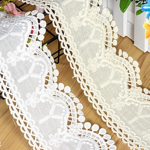 May include: White cotton lace trim with a scalloped edge and small pom-poms. The lace has a delicate floral pattern.
