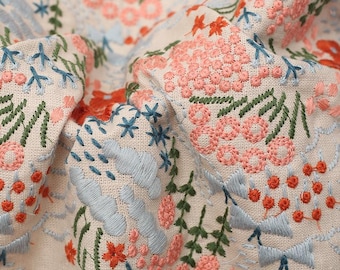 May include: Close-up of a beige fabric with embroidered floral designs in shades of pink, blue, and green. The fabric has a textured surface and a subtle sheen.