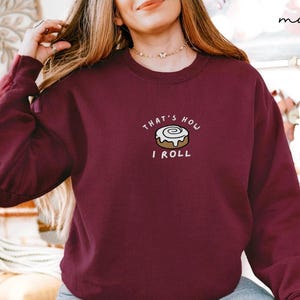 May include: Maroon sweatshirt with the text "THAT'S HOW I ROLL" and a graphic of a cinnamon roll. The long-sleeved crew neck top is a solid color. The sweatshirt is a casual style.