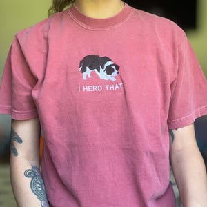 Puede incluir: Una camiseta de manga corta color rosa polvoriento con un bordado de un border collie blanco y negro en posici&oacute;n agachada. Debajo del perro, el texto "I HERD THAT" est&aacute; bordado en letras may&uacute;sculas blancas en el centro del pecho.