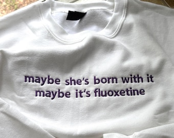 May include: A white crewneck sweatshirt featuring purple embroidered text on the chest that reads, maybe she's born with it maybe it's fluoxetine. The garment has long sleeves with ribbed cuffs and a ribbed neckline, constructed from a solid white fabric.