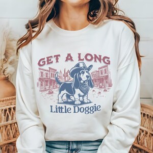 May include: A white long-sleeved sweatshirt with a graphic print of a cartoon basset hound wearing a cowboy hat and bandana. The text "Get a Long Little Doggie" is printed below the image.