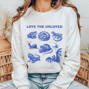 May include: White long-sleeve sweatshirt with the text "LOVE THE UNLOVED" in blue lettering. The sweatshirt features a blue graphic of a snake, frog, snail, mouse, spider, bat, and lizard.