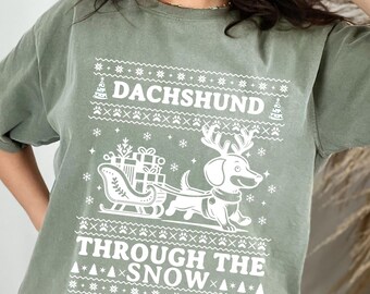 May include: A green t-shirt featuring a white dachshund design. The dachshund is pulling a sled with presents and the text "Dachshund Through The Snow" is printed below the image.