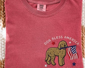 May include: Crimson-colored t-shirt with a graphic of a dog holding an American flag, with the text "DOG BLESS AMERICA" and "250TH ANNIVERSARY". The shirt is folded on a patterned surface.