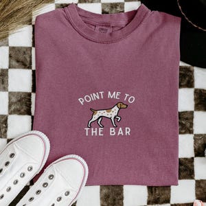 Pode incluir: Camiseta cor de berry com a frase bordada "Point Me To The Bar" e um gráfico de cachorro. A camiseta é combinada com tênis brancos e um chapéu preto. Enfeites decorativos de bolas de discoteca estão espalhados ao redor da camiseta.