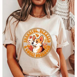 May include: A beige t-shirt with a graphic of a corgi dog and the text "Thankful, Blessed & Corgi Obsessed".